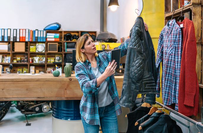 Blonde woman holding digital tablet while check leather jacket on second hand clothing store