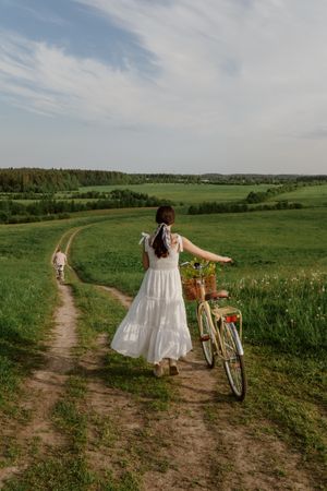 Back view of young woman in light dress walking with bicycle on green grass field