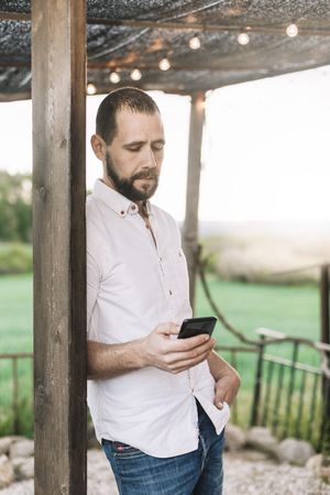 Bearded man standing at countryside home while using a mobile phone