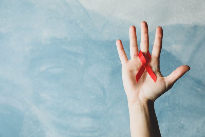 Hand and red awareness ribbon against blue background, space for text