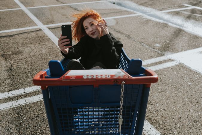 Smiling woman with smartphone in shopping trolley in parking lot