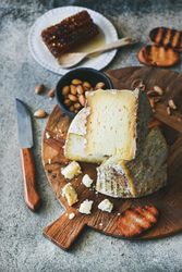 Wheel of cheese on wooden board, with knife, peanuts, crostini and honey