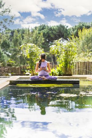 A woman holding a glass purple lotus while sitting in lotus pose on a tree  trunk over the water. Vertical shot