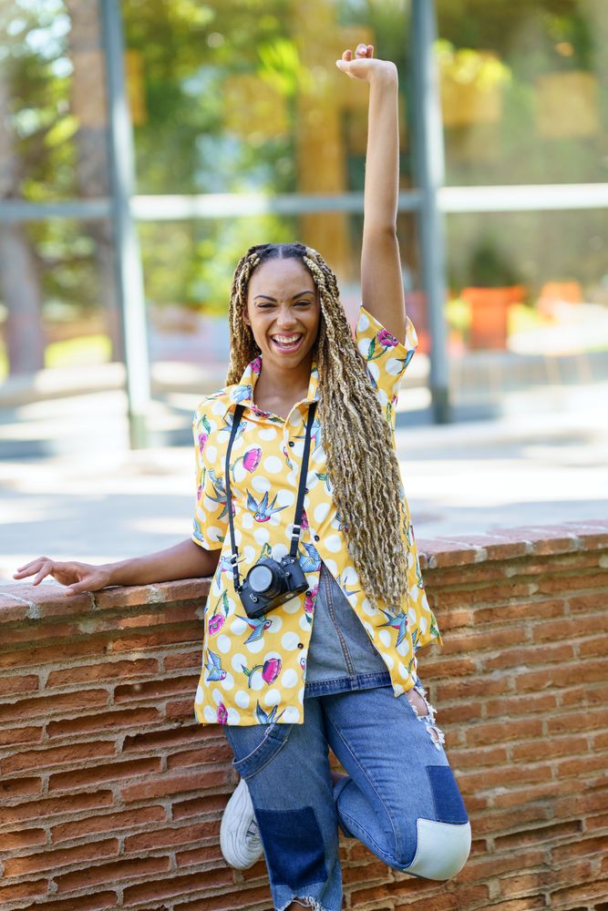 Joyous female in bold patterned shirt leaning on brick wall with camera