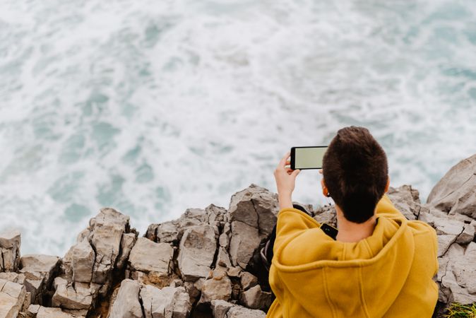 Woman in yellow hoodie taking selfie on smartphone on rocky shore