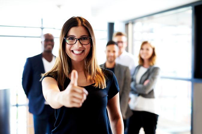 Businesswoman pictured in a bright office in front of her colleagues giving the thumbs up