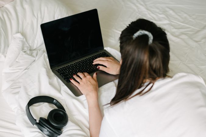 Millennial woman using laptop on bed with headphones