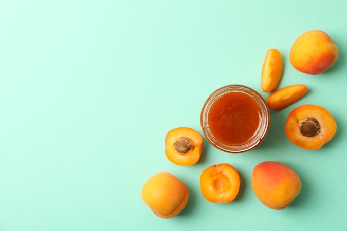 Glass jars with apricot jam and ingredients on mint background, top view