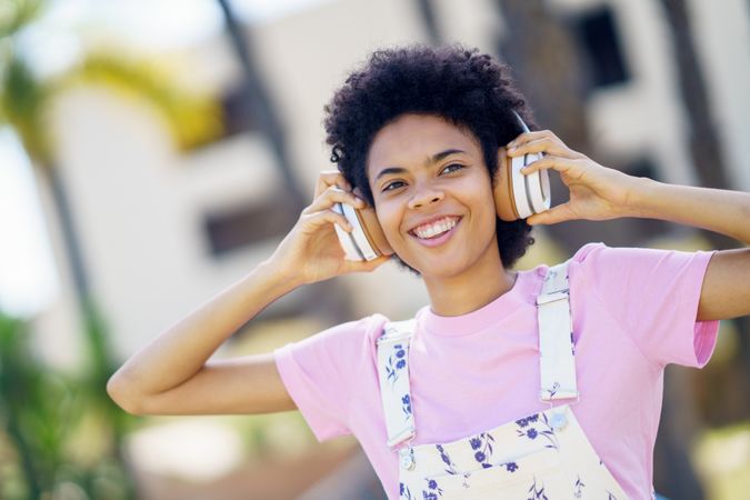 Happy female in floral overalls holding headphones over her ears in park