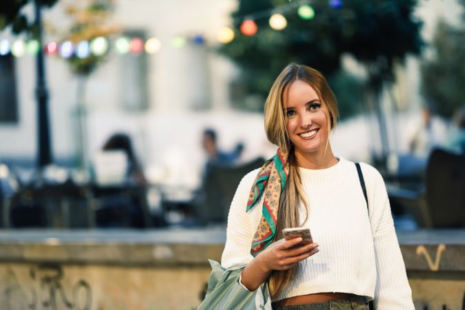Chic woman with scarf in hair smiling outside holding phone