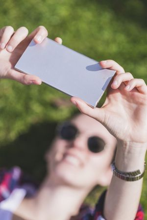 Above view of a young smiling hipster woman lying on grass in a sunny day at a park while taking a selfie with a mobile phone