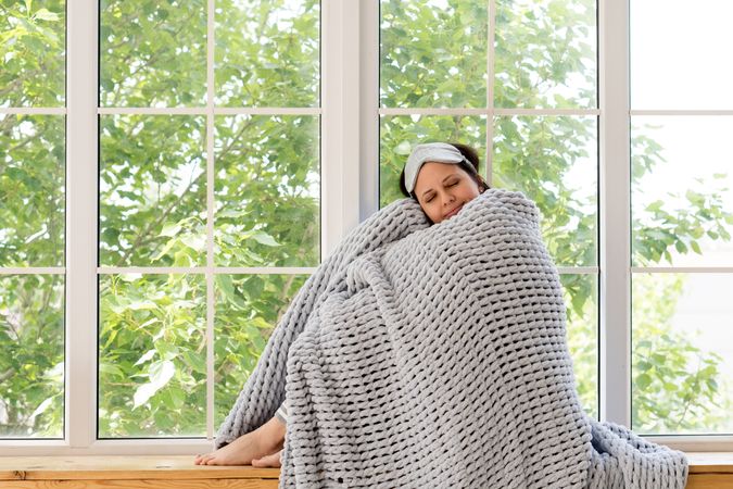 Woman sleeping, snuggled in chunky gray blanket by window.