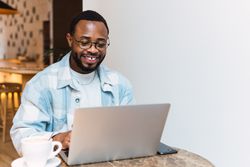 Delighted Black man with coffee browsing laptop
