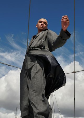 Stylish young man in belted gray coat blowing in the wind against a blue sky
