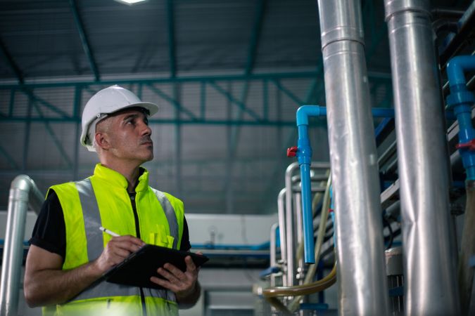A male industrial engineer wearing a safety helmet and reflective vest is inspecting equipment in a factory. He is using a digital tablet to review data. Background show industrial pipes and machinery