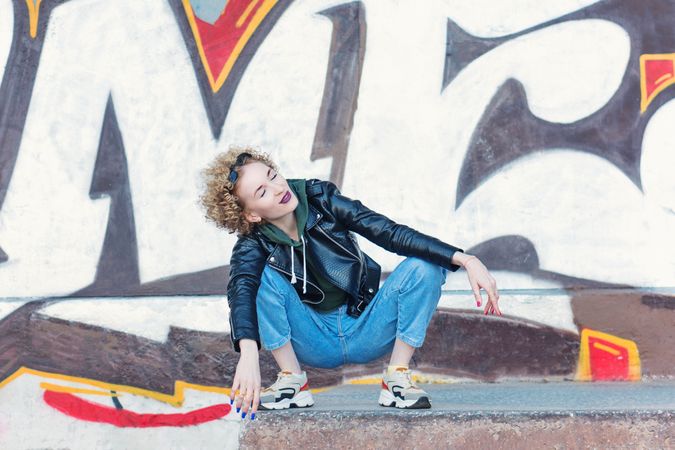 Full length portrait of trendy hip woman sitting at the graffiti wall background