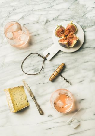 Two glasses of rose wine on marble table with quartered fig, cheese, knife, corkscrew