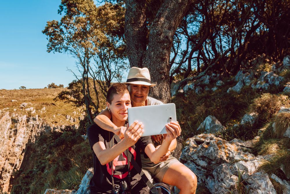 Older and younger woman looking at tablet sitting on stone in valley