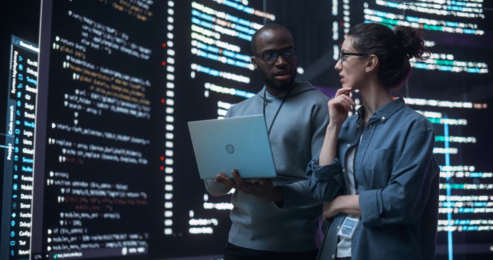 Portrait of Two Diverse Developers Using Laptop Computer, Discussing Lines of Code that Appear on Big Screens Surrounding Them.