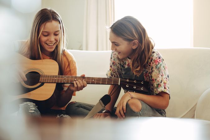 Girl learning to play guitar at home sitting with her friend