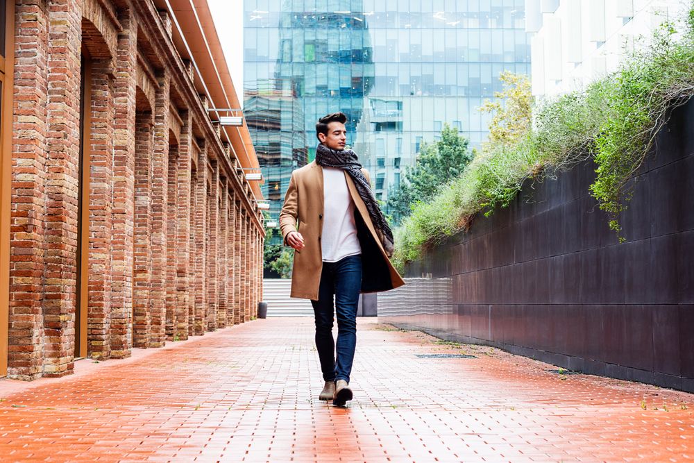 Man wearing camel coat and scarf looking around while walking down empty street