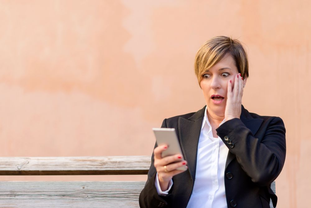 Surprised female in blazer sitting on bench checking phone in front of peach wall