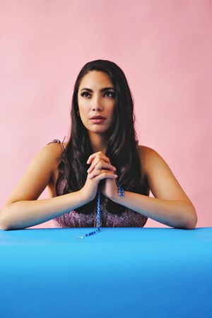 Hispanic woman looking away and praying with rosary beads in pink room, vertical