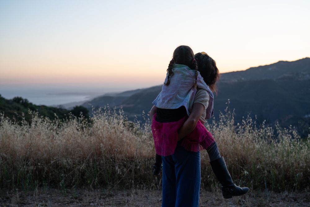 Back view of mom giving piggy back to her daughter and looking toward view of mountains and ocean