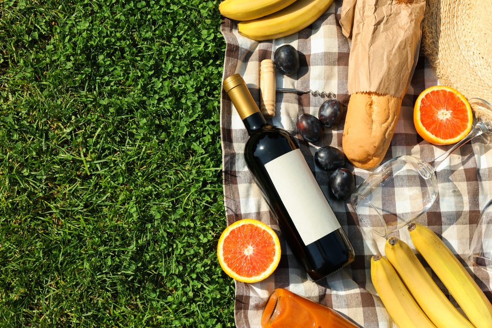 Bottle of wine, baguette, fruits, straw hat and towel on grass, space for text