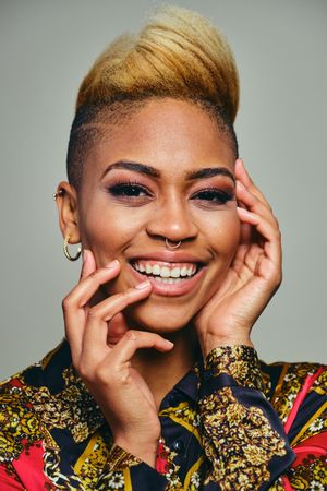 Portrait of happy Black woman with short blonde hair in bold patterned shirt and hands to her face