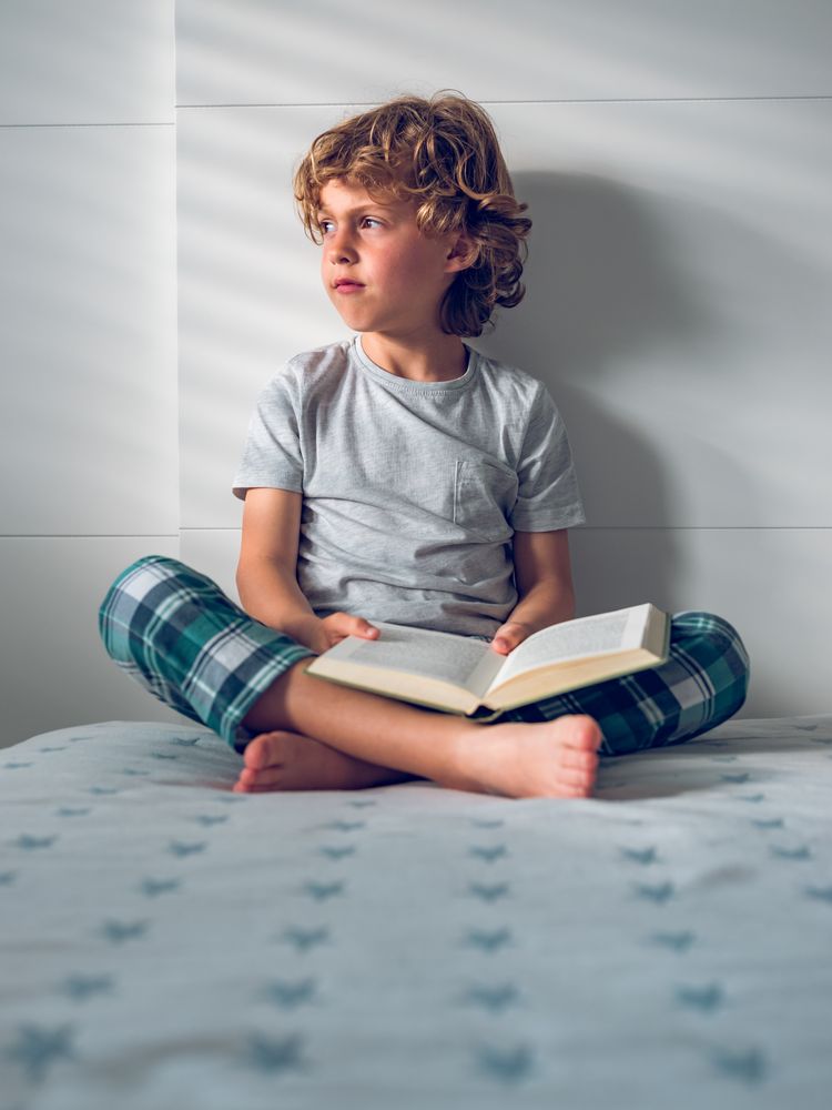 Pensive little boy sitting on bed with opened book