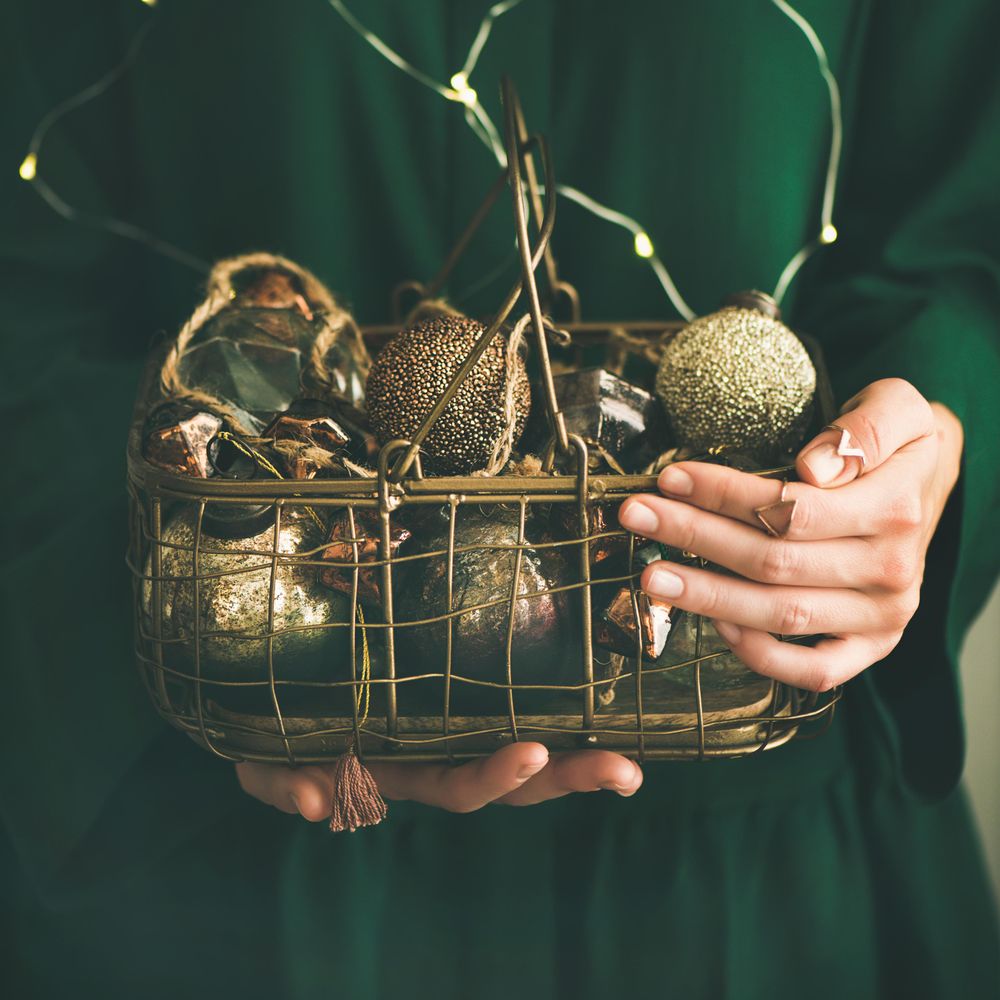 Woman in green dress holding wire basket of gold holiday decorations, square crop