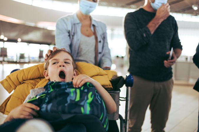 Boy yawning while sitting on luggage trolley at airport with parents in background