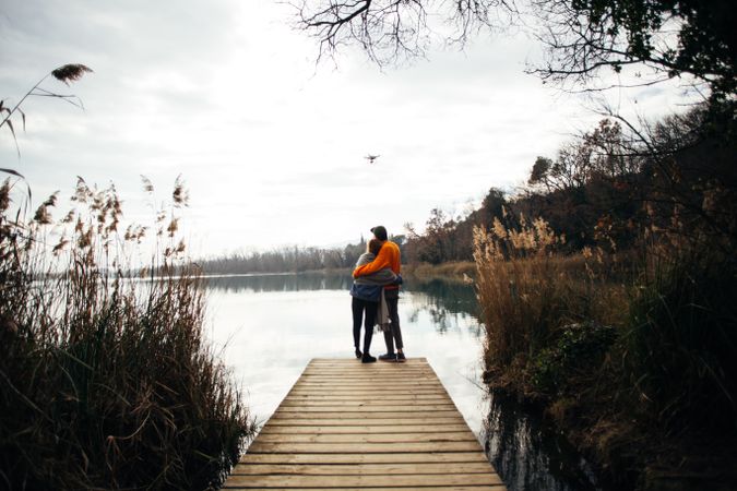 Man and woman embrace on the pier of a lake looking up at a drone