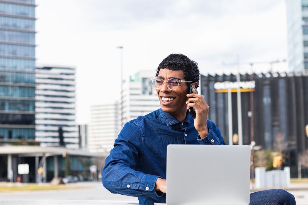 Happy businessman freelancer working on laptop and talking on mobile phone in city