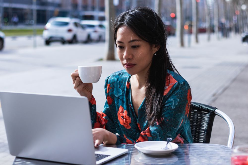 Long-haired brunette Asian woman having a coffee on a terrace of a cafe while is working with a laptop