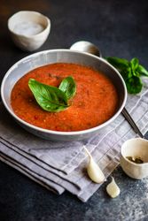 Grey bowl of gazpacho soup with basil leaves on napkin served with seasoning