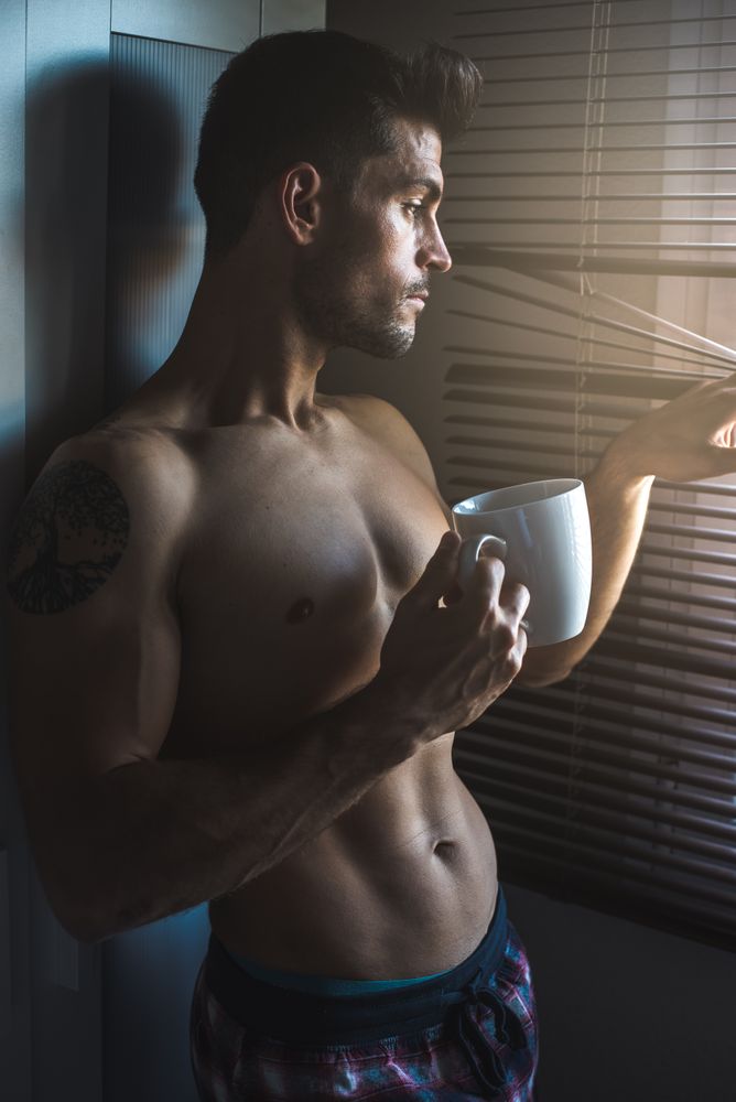 Man standing by a windows blind with a cup of coffee