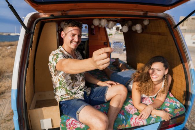 Smiling man raising preservative in hand sitting with woman in trunk of car