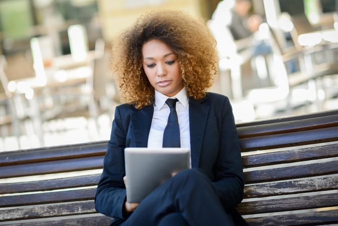 Woman in suit intently reading something on her digital tablet’s screen on outdoor bench