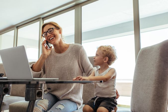 Mother working from home with laptop and talking on cell phone with son sitting nearby