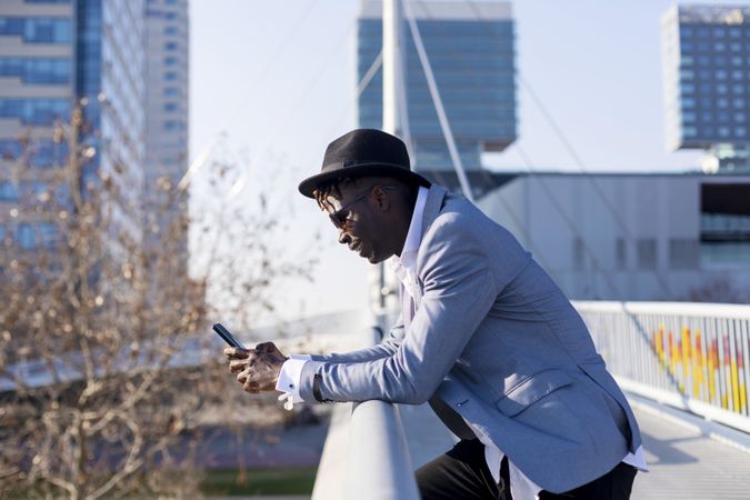 Side view of Black man wearing hat and sunglasses leaning on a metallic fence while using a mobile phone outdoors in a sunny day