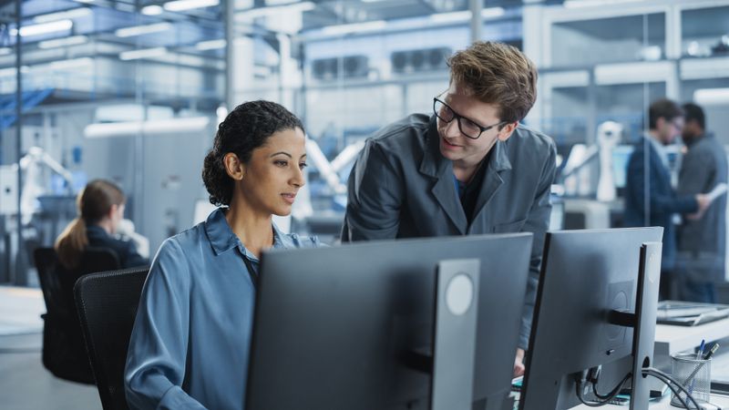 Portrait of Young Male Assembly Line Worker and Hispanic Female Engineer Collaborating on a Project, Using Computer and Talking in a Factory with Robotic Arms Producing Devices for Tech Industries.