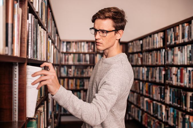 Male college student picking book from library wall