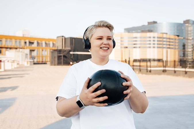 Smiling blonde woman with medicine ball, smart watch and headphones on a rooftop