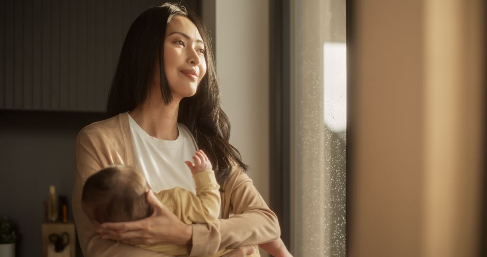 Portrait of an Asian Mother Holding her Baby in her Arms and Looking at the Street Through a Big Window at Home.
