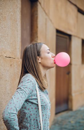 Side view of woman standing outside in front of stone wall blowing bubble gum