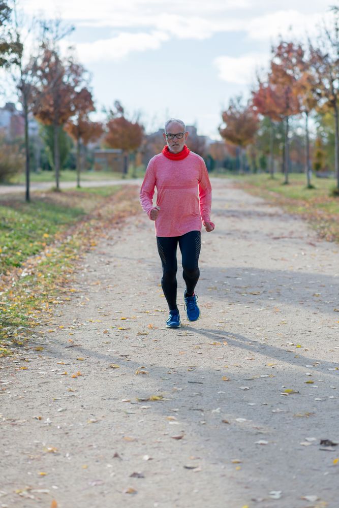 Grey haired man in red shirt jogging on in tree lined park