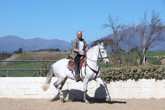 Man in checkered shirt riding horse on sandy ground around paddock