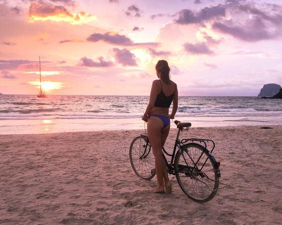 Woman wearing biking standing beside bicycle on the beach at sunset